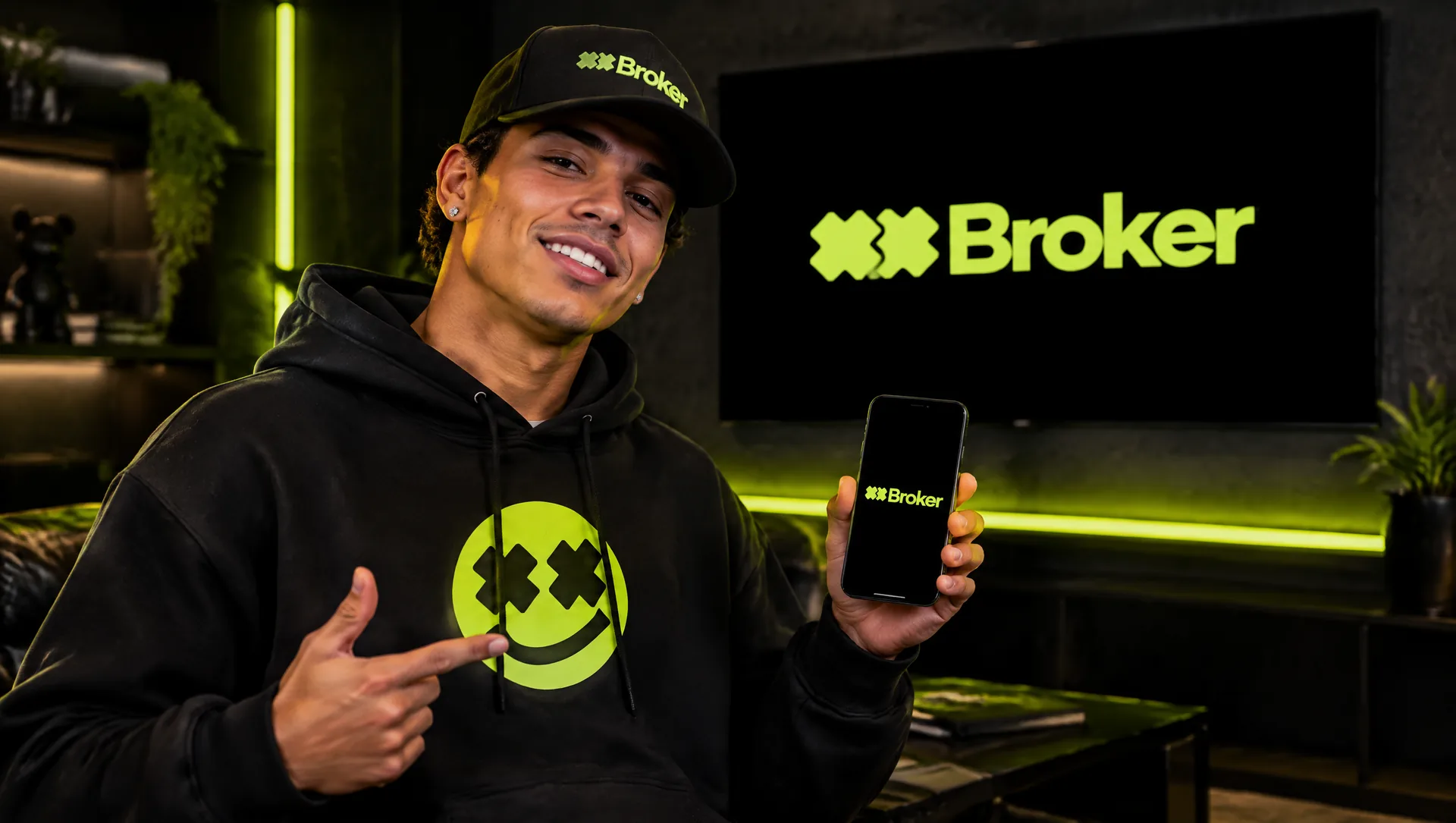 Pessoa de moletom e boné XXBroker apontando o celular para a tela com a logo da marca